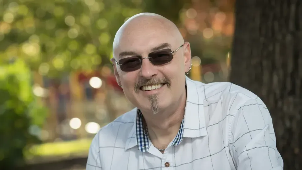 Mark Shannon, smiling, outdoors under a tree in dappled light